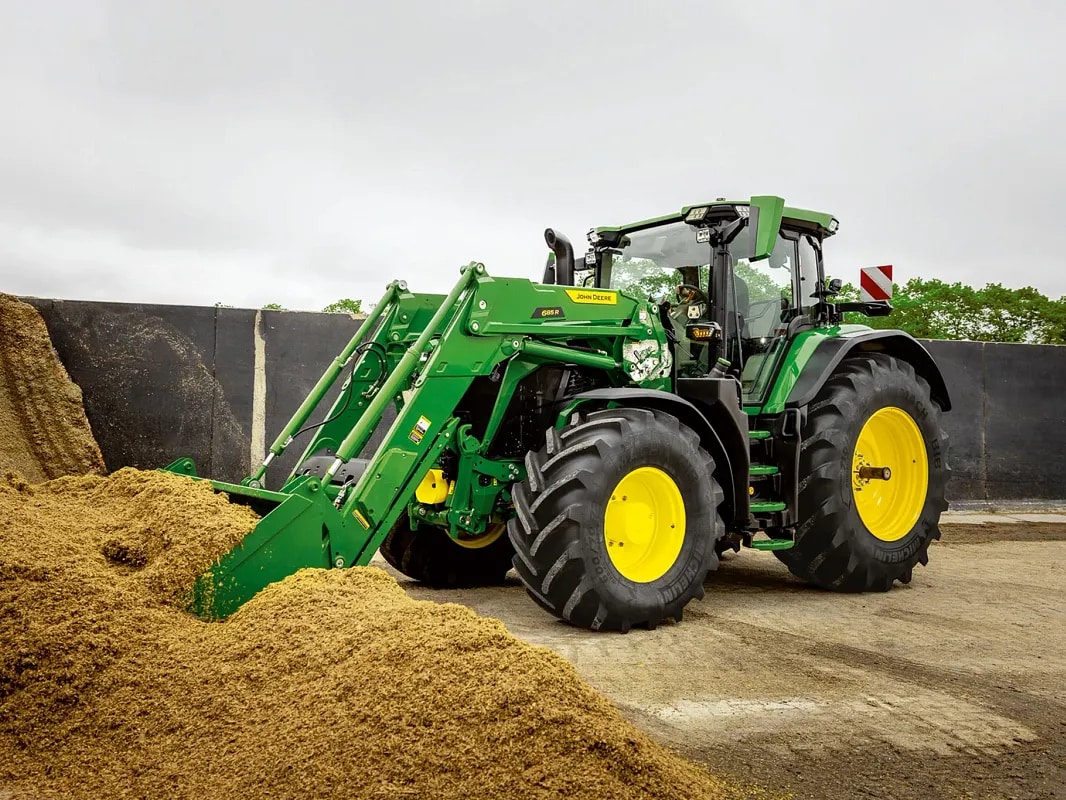 טרקטור John Deere דוחף לערימת מספוא באמצעות כף מעמיס קדמי בסביבת חווה.