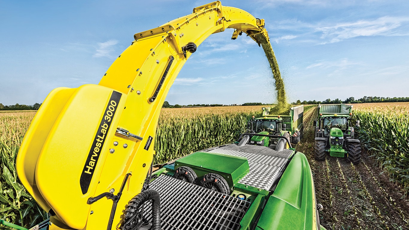 מבט מאחור על מקצרת מספוא ממונעת סדרת John Deere 9000 בעלת שתי עגלות מספוא הנגררות ע&quot;י טרקטורי John Deere ישר מאחור