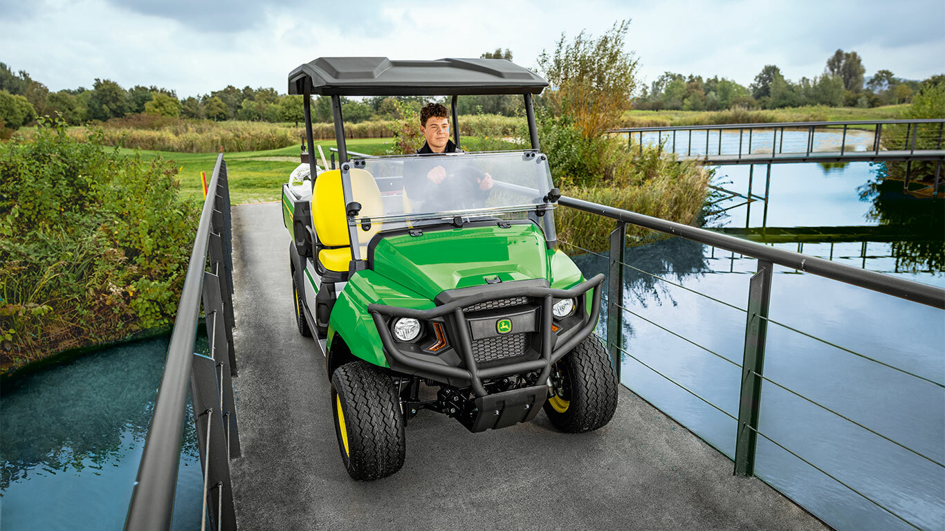 רכב ייעודי John Deere Gator GS נוסע לאורך גשר צר מעל תעלת מים שקטים, מוקף בצמחייה שופעת ועצים.