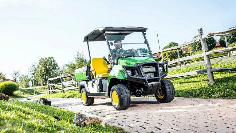 רכב ייעודי ירוק John Deere Gator GS נוסע בכביש סלול דרך שטח שעבר גינון עם דשא וגדרות עץ.