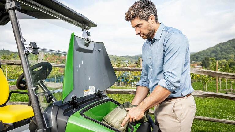 אדם המניח תיק מרופד בתא האחסון הקדמי של John Deere Gator GS ירוק שחונה בחוץ ליד גדר עץ וכרם.