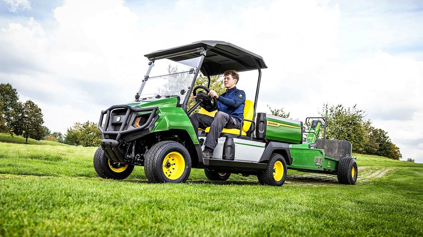 רכב ייעודי John Deere Gator חשמלי ירוק גורר ציוד בשדה עשבים תחת שמיים מעוננים חלקית.