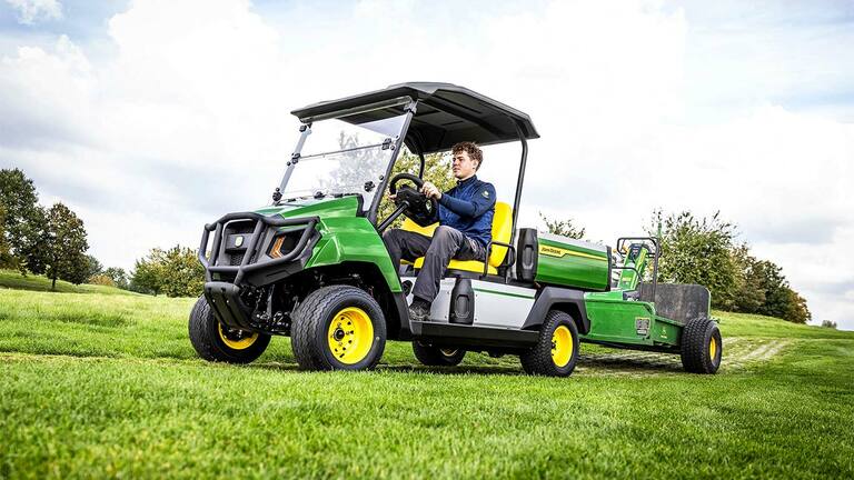 רכב ייעודי John Deere Gator חשמלי ירוק גורר ציוד בשדה עשבים תחת שמיים מעוננים חלקית.
