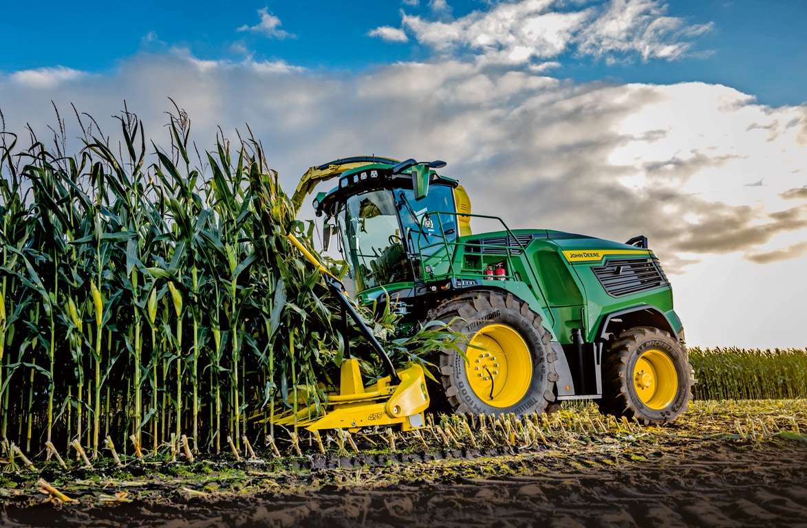 מקצרת מספוא John Deere קוצרת תירס בשדה תחת שמיים כחולים עם עננים.