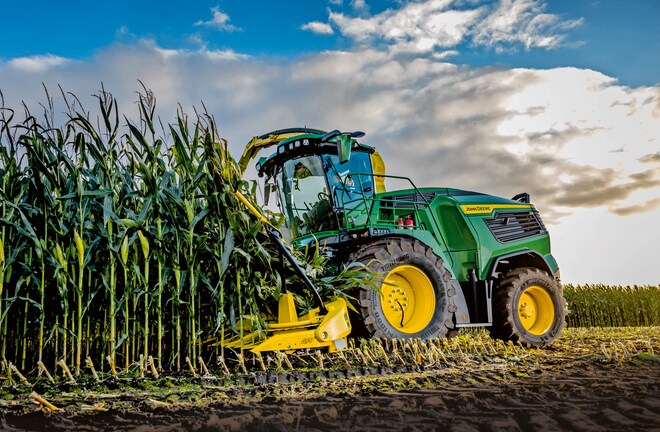 מקצרת מספוא John Deere קוצרת תירס בשדה תחת שמיים כחולים עם עננים.
