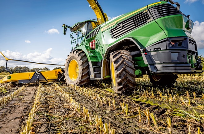 מקצרת מספוא של John Deere עובדת בשדה תירס שנקצר, ומציגה את הצמיגים הגדולים של המכונה ואת מנגנון החיתוך מול שמים כחולים ובהירים.