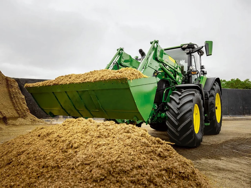 טרקטור John Deere משתמש במעמיס קדמי לנשיאת תחמיץ קצוץ באזור אחסון חקלאי.